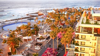 A tropical coastal resort with numerous palm trees, multiple swimming pools, and people walking along pathways. The image includes a large building with balconies and outdoor seating areas on the right. The ocean with waves crashing against rocks is visible in the background.