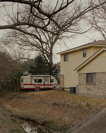 A parked travel trailer is next to a house with yellow siding and brick. Bare tree branches stretch across the sky, indicating it might be winter. The ground is covered with dry leaves, and there's a small drainage ditch in the foreground.