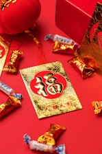 Red and gold decorations and candies are arranged on a red surface. A gold card with Chinese characters and floral patterns is prominently displayed, surrounded by candy wrappers with vibrant designs. A red lantern and a decorative box also feature prominently, contributing to a festive atmosphere.