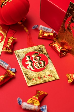 Red and gold decorations and candies are arranged on a red surface. A gold card with Chinese characters and floral patterns is prominently displayed, surrounded by candy wrappers with vibrant designs. A red lantern and a decorative box also feature prominently, contributing to a festive atmosphere.