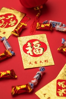 A collection of red and gold envelopes with traditional Chinese characters are arranged on a red surface, accompanied by assorted candies. A red tassel and a part of a traditional lantern are visible, enhancing the festive theme.