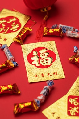A collection of red and gold envelopes with traditional Chinese characters are arranged on a red surface, accompanied by assorted candies. A red tassel and a part of a traditional lantern are visible, enhancing the festive theme.