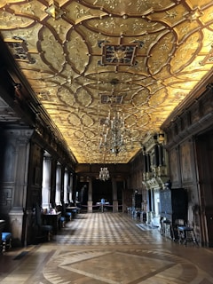 An opulent and expansive interior room featuring an intricately designed golden ceiling with ornate patterns and chandeliers. The walls are adorned with dark wood paneling, and the parquet floor is intricately patterned. The room is lined with chairs and tables, and there is a sense of grandeur and luxury.