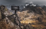 A rugged mountain landscape with a focus on a flexible tripod holding a camera or phone, positioned on a rocky surface. The background features majestic mountains partially covered in snow under a moody, overcast sky.