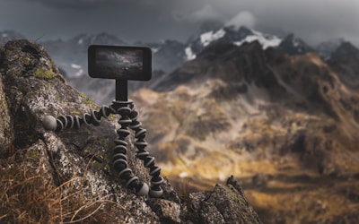 A rugged mountain landscape with a focus on a flexible tripod holding a camera or phone, positioned on a rocky surface. The background features majestic mountains partially covered in snow under a moody, overcast sky.