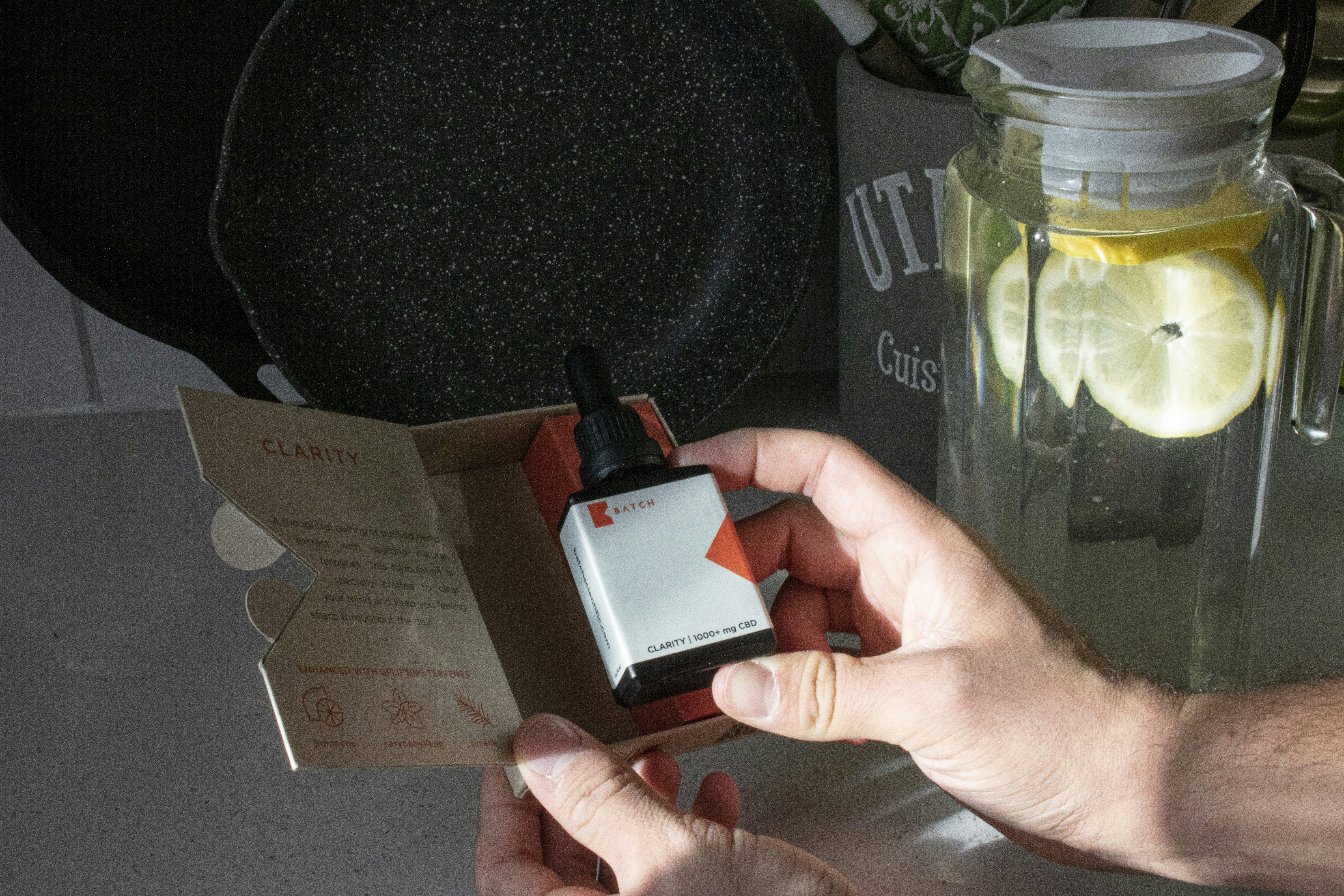 Male hand holding CBD oil tincture in front of kitchenware and countertop with light rays refracting through lemon water container in the morning
