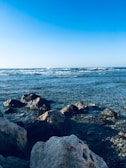 A serene ocean view with gentle waves lapping against large rocks along the shore. The sky is clear and blue, adding to the peaceful atmosphere.