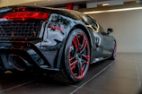 A sleek black sports car is shown from the rear side, emphasizing its polished finish and aerodynamic design. The car features red accents around the taillights and red detailing on the rims of its low-profile tires. The indoors showroom setting is clean and modern with lighting that highlights the car's glossy surface.
