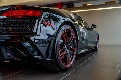 A sleek black sports car is shown from the rear side, emphasizing its polished finish and aerodynamic design. The car features red accents around the taillights and red detailing on the rims of its low-profile tires. The indoors showroom setting is clean and modern with lighting that highlights the car's glossy surface.
