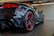 A sleek black sports car is shown from the rear side, emphasizing its polished finish and aerodynamic design. The car features red accents around the taillights and red detailing on the rims of its low-profile tires. The indoors showroom setting is clean and modern with lighting that highlights the car's glossy surface.