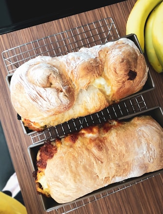 Two loaves of freshly baked bread sit in metal trays on a wire cooling rack atop a wooden surface. The bread has a golden-brown crust with some flour dusting on the surface. A bunch of bananas is visible on the right side of the image.