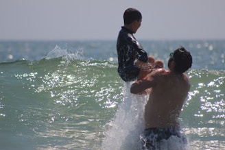 A man is playfully lifting a child above the waves in the ocean. The water around them is sparkling under the sunlight, and the ocean stretches out to the horizon.