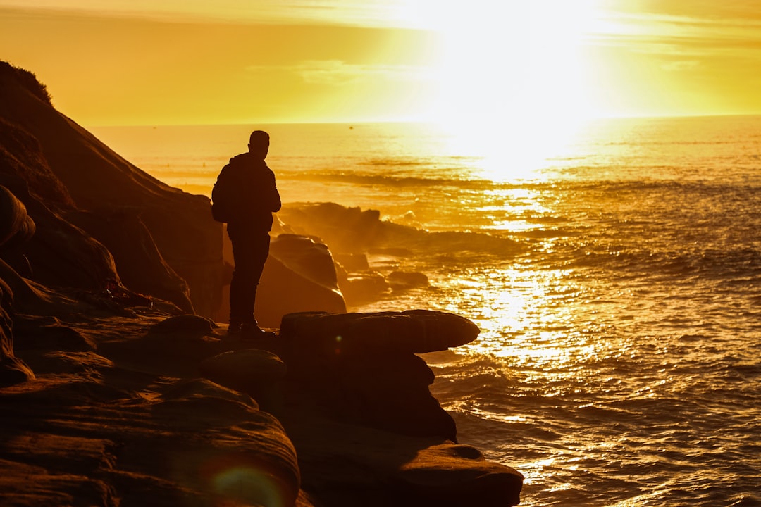 silhouette of man standing on rock formation near body of water during sunset