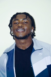 A man with short, braided hair and a subtle smile wears a two-toned Nike jacket and black shirt. He has earbuds in both ears against a plain light background.