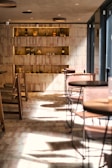 A cozy, warmly-lit cafe interior with wooden furniture and shelves. The shelves on the back wall display various ceramic and glass pots and jars, adding a rustic charm. Natural light creates interesting shadow patterns on the floor, enhancing the serene and inviting atmosphere.