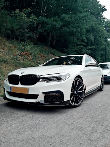 A sleek white car parked on a roadside with lush green trees in the background. The vehicle's shiny surface and modern design are highlighted by its surroundings, with large black alloy wheels and signature branding.