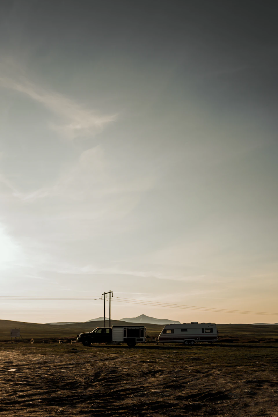 Two vehicles parked on a dirt road at sunset amidst rural landscape with mountains