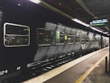 A black passenger train is stationed at a platform inside a train station, with fluorescent lights illuminating the scene. The side of the train has large windows and the platform is marked with yellow lines. There is a digital sign board above displaying destinations and times.