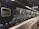 A black passenger train is stationed at a platform inside a train station, with fluorescent lights illuminating the scene. The side of the train has large windows and the platform is marked with yellow lines. There is a digital sign board above displaying destinations and times.