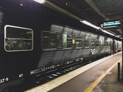A black passenger train is stationed at a platform inside a train station, with fluorescent lights illuminating the scene. The side of the train has large windows and the platform is marked with yellow lines. There is a digital sign board above displaying destinations and times.