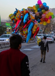 A street scene with a person walking away while holding a large bunch of colorful balloons shaped like various characters, including superheroes and animals. Another person is visible in the foreground, partially facing the balloon vendor. Cars line the street, and there are buildings and neon signs in the background.