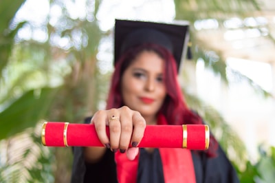 A person in a graduation cap and gown holds a red diploma case towards the camera. The background is blurred, featuring green foliage.