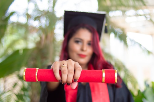 A person in a graduation cap and gown holds a red diploma case towards the camera. The background is blurred, featuring green foliage.
