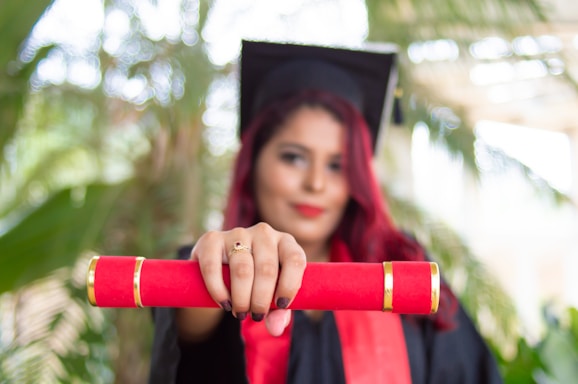 A person in a graduation cap and gown holds a red diploma case towards the camera. The background is blurred, featuring green foliage.