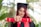 A person in a graduation cap and gown holds a red diploma case towards the camera. The background is blurred, featuring green foliage.