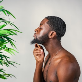 A man is grooming his beard with a comb, standing in profile against a plain background. A lush green plant is visible on the left side of the image, adding a natural element to the scene. The man's skin tone is complemented by the neutral backdrop and the greenery.