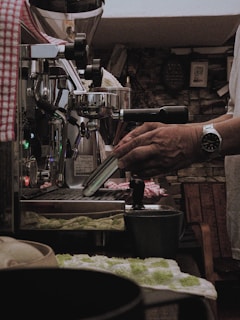 A person is operating an espresso machine in a cozy, dimly lit setting. The machine has metal parts and a lever, with a hand operating it. A cup and a green and white towel are visible on the counter, alongside various other small objects. The background consists of a rustic stone wall with some pictures or decorations.