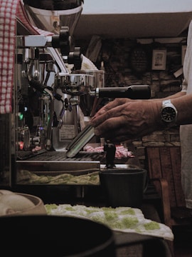 A person is operating an espresso machine in a cozy, dimly lit setting. The machine has metal parts and a lever, with a hand operating it. A cup and a green and white towel are visible on the counter, alongside various other small objects. The background consists of a rustic stone wall with some pictures or decorations.