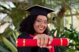 A person in a graduation cap and gown is smiling while holding a red diploma. The background is filled with lush green plants, giving a vibrant and celebratory atmosphere. The individual is wearing glasses and has curly hair.