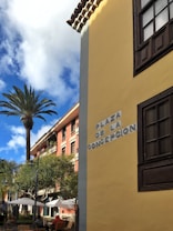 An outdoor scene with a tall palm tree and a pastel-colored building with a sign reading 'Plaza de la Concepcion'. The corner of another building with a yellow wall and dark wooden window frames is visible in the foreground. Beneath the trees, there are tables with umbrellas and people seated, suggesting a caf&eacute; or outdoor dining setting. The sky is partly cloudy, providing a bright and sunny atmosphere.