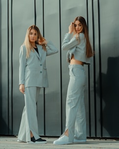 Two women stand in front of a dark, vertical paneled wall. Both are wearing light-colored suits with wide-legged pants. The woman on the left has her arm raised, touching her hair, and looking directly at the camera. The woman on the right is adjusting her hair with one hand and looking down. They both have long, straight hair and are wearing casual footwear.