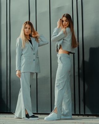Two women stand in front of a dark, vertical paneled wall. Both are wearing light-colored suits with wide-legged pants. The woman on the left has her arm raised, touching her hair, and looking directly at the camera. The woman on the right is adjusting her hair with one hand and looking down. They both have long, straight hair and are wearing casual footwear.