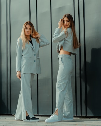 Two women stand in front of a dark, vertical paneled wall. Both are wearing light-colored suits with wide-legged pants. The woman on the left has her arm raised, touching her hair, and looking directly at the camera. The woman on the right is adjusting her hair with one hand and looking down. They both have long, straight hair and are wearing casual footwear.