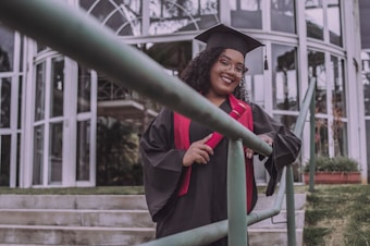 A person wearing a graduation cap and gown is standing on outdoor stairs, holding a diploma or certificate. The background features glass windows and a railing in the foreground.