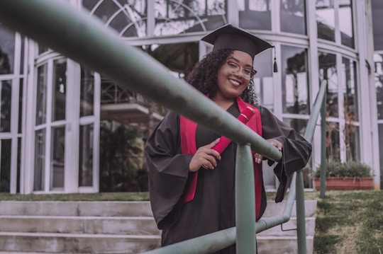 A person wearing a graduation cap and gown is standing on outdoor stairs, holding a diploma or certificate. The background features glass windows and a railing in the foreground.