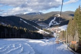 A scenic mountain landscape features a ski resort with snow-covered slopes and tall evergreen trees. Chairlifts are visible transporting skiers up the mountain, and clusters of buildings, possibly lodges, are nestled in the valley below. The sky is partly cloudy, and the distant peaks are shrouded in snow.