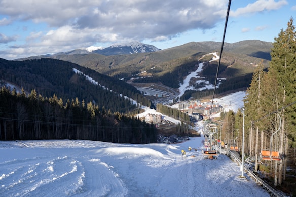 A scenic mountain landscape features a ski resort with snow-covered slopes and tall evergreen trees. Chairlifts are visible transporting skiers up the mountain, and clusters of buildings, possibly lodges, are nestled in the valley below. The sky is partly cloudy, and the distant peaks are shrouded in snow.