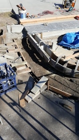 A construction site with concrete forms and wooden planks arranged on the ground. Tools and materials are scattered around, including a blue tarp, bucket, cutting tools, and electrical cables. Sunlight casts shadows on the concrete surface.