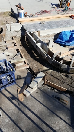 A construction site with concrete forms and wooden planks arranged on the ground. Tools and materials are scattered around, including a blue tarp, bucket, cutting tools, and electrical cables. Sunlight casts shadows on the concrete surface.