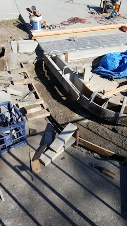 A construction site with concrete forms and wooden planks arranged on the ground. Tools and materials are scattered around, including a blue tarp, bucket, cutting tools, and electrical cables. Sunlight casts shadows on the concrete surface.
