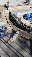 A construction site with concrete forms and wooden planks arranged on the ground. Tools and materials are scattered around, including a blue tarp, bucket, cutting tools, and electrical cables. Sunlight casts shadows on the concrete surface.
