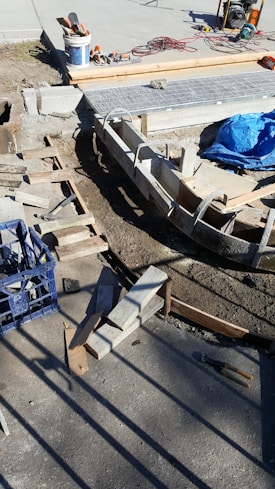 A construction site with concrete forms and wooden planks arranged on the ground. Tools and materials are scattered around, including a blue tarp, bucket, cutting tools, and electrical cables. Sunlight casts shadows on the concrete surface.