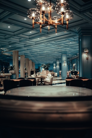 An elegant dining area with a sophisticated design, featuring plush chairs and round tables set against a backdrop of ornate columns and a coffered ceiling. The warm lighting from the chandelier enhances the luxurious ambiance.