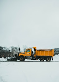 A yellow snowplow truck is parked on a snow-covered road with bare trees in the background. The truck is equipped with a large plow at the front and is filled with sand or salt in its dump bed for de-icing purposes. The sky is overcast, and the landscape is dominated by the stark white of the snow.