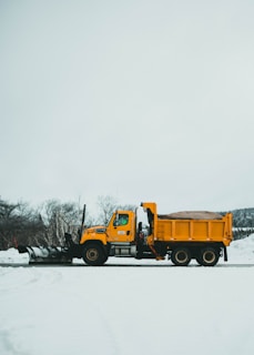 A yellow snowplow truck is parked on a snow-covered road with bare trees in the background. The truck is equipped with a large plow at the front and is filled with sand or salt in its dump bed for de-icing purposes. The sky is overcast, and the landscape is dominated by the stark white of the snow.