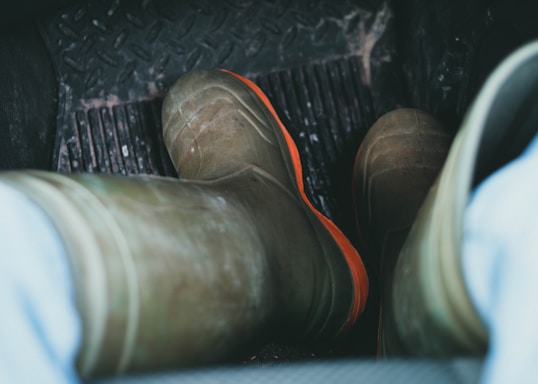 View of a pair of worn work boots with orange soles, placed on the rubber floor mat of a vehicle interior. The floor mat has a rugged texture, and the surrounding floor area seems to be somewhat dirty, indicating use in muddy or rough environments.